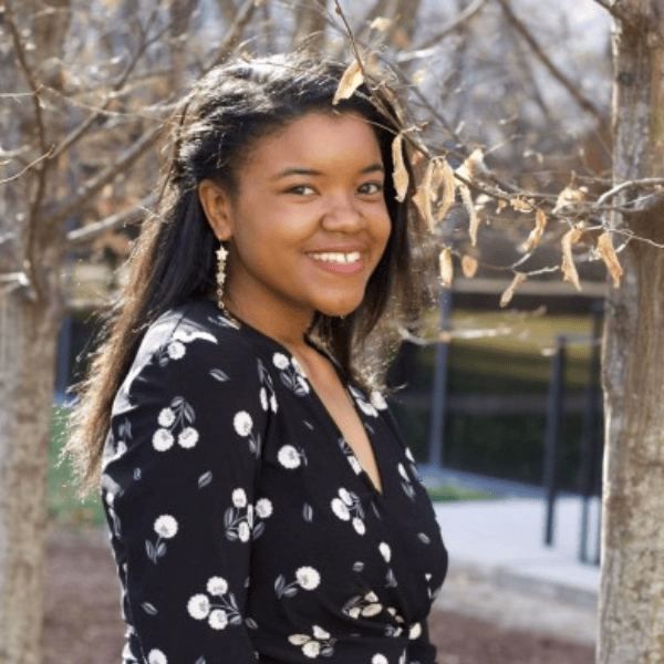 Woman smiling outdoors in fall, wearing a black floral dress, surrounded by trees.