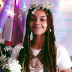 Young woman with braided hair and floral crown smiling at the camera during an indoor event.