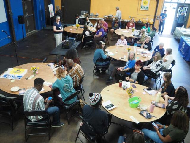 Networking event with diverse community members, some engaging with phones, others chatting, in a lively indoor setting.