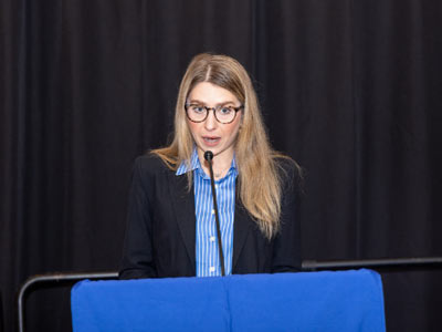 Woman speaking at Michigan Climate Action Network event with microphone and black curtain background.