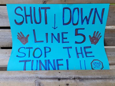 Protest sign reading 'Shut Down Line 5 Stop the Tunnel' with handprints and stop sign.