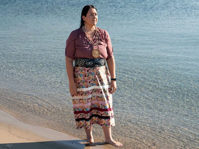 Native woman standing by the water with traditional clothing and jewelry.